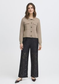 Cardigan corto beige con colletto e due tasche frontali, abbinato a pantaloni palazzo neri in pizzo con intricati motivi a taglio.