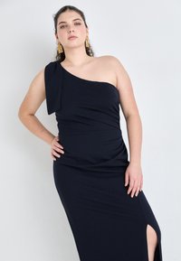 Navy blue, one-shoulder dress made of stretch fabric featuring a ruched texture and a thigh-high slit, accessorized with gold earrings.