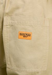 Khaki fabric with a textured finish; features a patch label in bright orange reading "Service Works," located on the pocket.