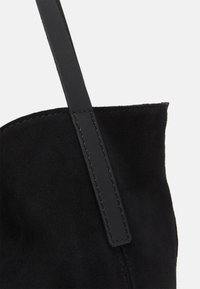 Coin d'un sac en daim noir avec un détail de sangle en cuir cousue sur un fond blanc.