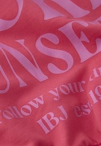 Koraltyg med stora rosa bokstäver. Texten inkluderar "FÖLJ DINA DRÖMMAR" och "IBJ est. 10" i olika typsnittsstorlekar. Slät textur.