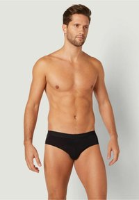 Schwarze Baumwollslips mit nahtlosem Design und elastischem Bund, die eine enge Passform und eine glatte Textur bieten, getragen von einem stehenden männlichen Model.