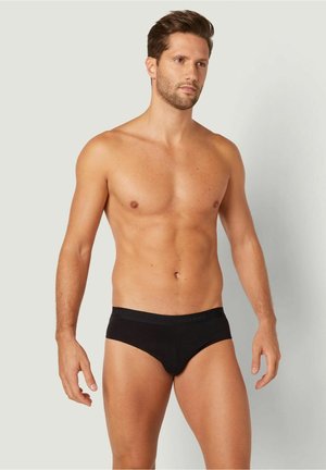 Schwarze Baumwollslips mit nahtlosem Design und elastischem Bund, die eine enge Passform und eine glatte Textur bieten, getragen von einem stehenden männlichen Model.
