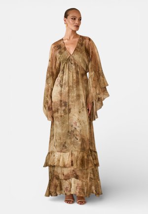 Donna che indossa un abito lungo e fluente in tonalità terra con maniche larghe, scollatura a V e orlo arricciato, in piedi davanti a uno sfondo semplice.