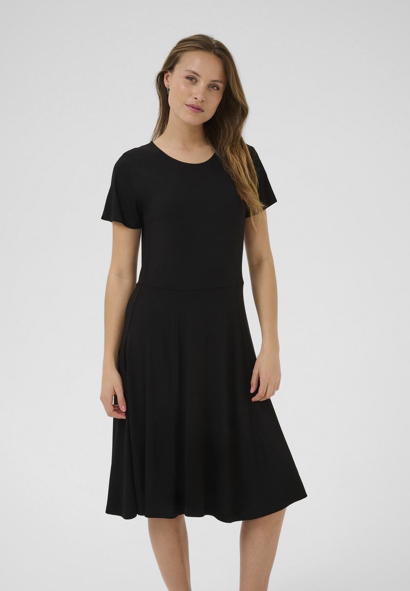 Femme aux longs cheveux portant une robe noire à manches courtes, longueur genoux, debout devant un fond clair uni.