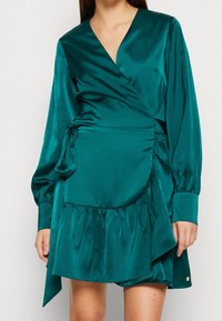 Tyrkysové saténové šaty s zavinovacím designem, dlouhými rukávy, hlubokým výstřihem do V a volánkovým lemem. Obsahují boční zavázání a hladkou texturu. Zlaté kovové akcenty.