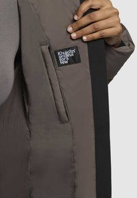 Pehmendatud jope muted pruunis, külgtasku ja musta aktsentriba. Silt ütleb "KhujooTm originaal mõne jaoks." Sile tekstuur.