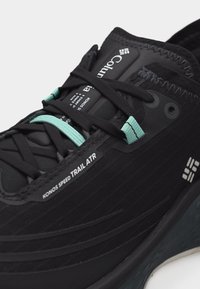 Κοντινό πλάνο μαύρου παπουτσιού για τρέξιμο σε μονοπάτι με μαύρα και γαλάζια κορδόνια, ετικέτα "KONOS SPEED TRAIL ATR" και λογότυπο Columbia στη γλώσσα και στην πλευρά.