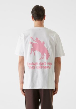 Hvit bomull T-skjorte med en rosa grafikk av en cowboy på hest med teksten "Cowboys tar ikke lur. De får i seg koffein." på baksiden.