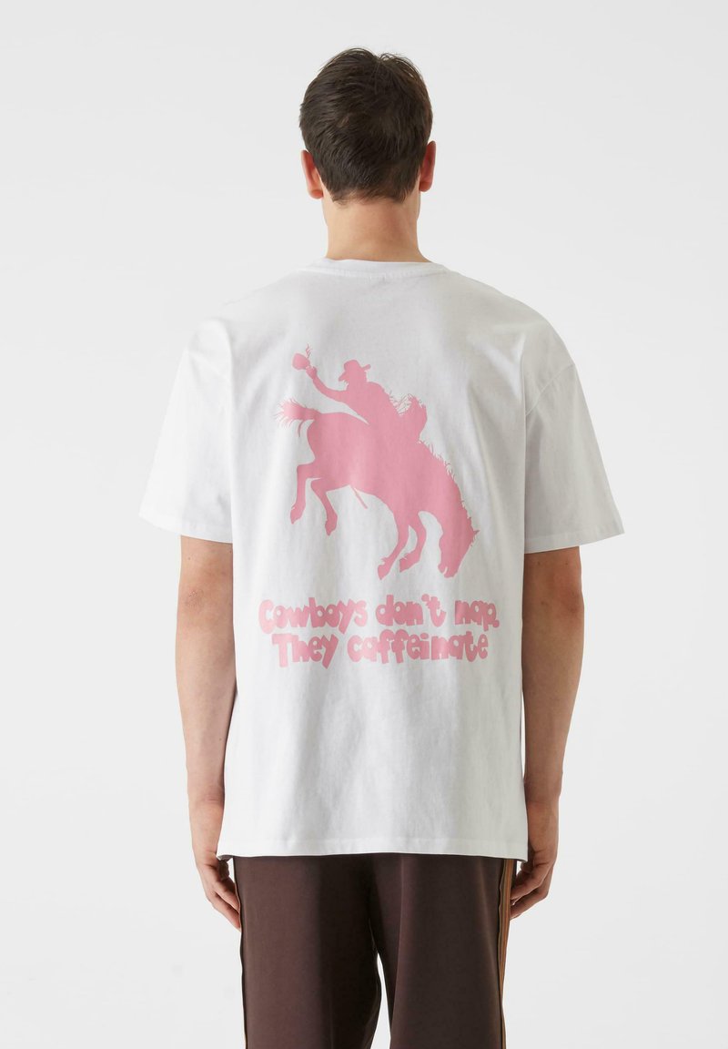Biela bavlnená tričko s ružovým grafickým zobrazením kovboja na koni a textom "Kovboji nespia. Kofeinizujú." na zadnej strane.