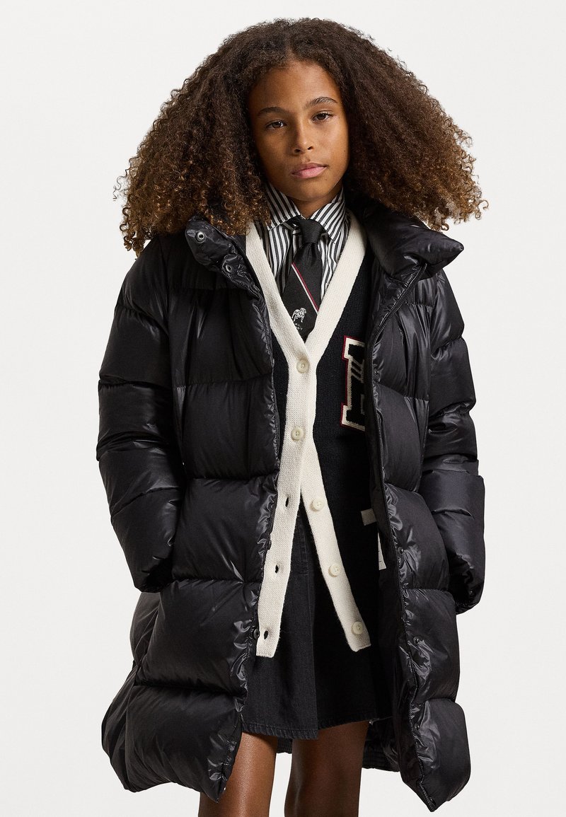 Fekete puffernadrág fényes felülettel, cipzáros záródással, magas gallérral és folt zsebekkel, egy krém színű kardigán és fekete ruha fölött viselve.