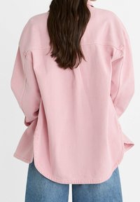 Camisa de mezclilla rosa con un corte relajado, que presenta mangas largas, un dobladillo redondeado y costuras visibles. Combinada con jeans de mezclilla azul claro.