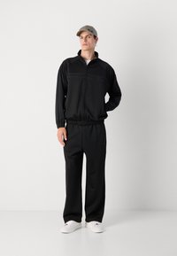 Fato de treino preto com sweat jacket com fecho éclair, apresentando acabamentos texturizados e calças com um cós elástico; usado com um chapéu de cor clara e ténis.