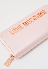 Roza sintetična usnjena denarnica z dvignjenimi metalnimi črkami, ki ustvarjajo napis "LOVE MOSCHINO", z zlatim zadrgo, okrašenim s srcem.