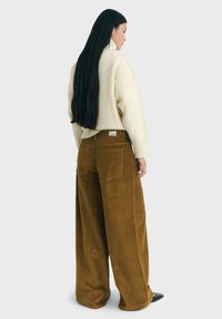Woman with long black hair wearing a cream oversized turtleneck sweater and wide-leg brown corduroy pants, standing sideways on white background.
