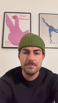 Gorro de malha canelada verde, usado na cabeça, com um suéter escuro. O fundo apresenta duas obras de arte emolduradas: uma gansa rosa e uma figura azul.