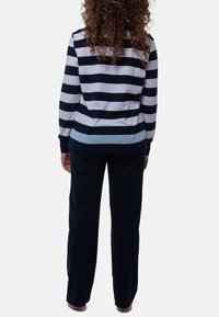 Person med lockigt hår som bär en långärmad marinblå och vit randig tröja och enfärgade marinblå byxor, stående barfota med ryggen vänd mot kameran.