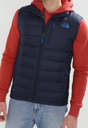 Mand iført navyblåt polstret vest over rød hættetrøje og lyseblå jeans mod en ensfarvet baggrund.