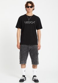 Black cotton t-shirt with white "Volcom" logo, paired with gray denim cargo shorts. Model wears black sunglasses and white and black socks.