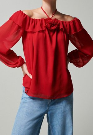 Femme portant un chemisier rouge transparent à épaules dénudées avec bordure à volants et détail fleur, assorti à un jean bleu clair, mains posées sur les hanches.