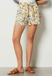 White shorts with a lemon and leaf pattern, featuring a gathered waistband and side pockets. Paired with brown leather slide sandals.