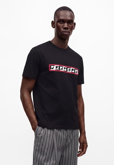 Homme portant un t-shirt noir avec le logo "VERSACE" et un pantalon gris rayé, debout devant un fond blanc uni.
