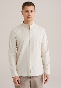 Camisa de manga larga color beige claro con cuello de banda, cierre frontal de botones y un ajuste relajado, hecha de una tela suave y texturizada.