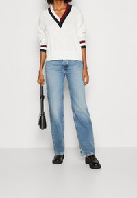 Weißer Pullover mit V-Ausschnitt und navyblauen sowie weinroten gestreiften Bündchen, kombiniert mit hellblauen Jeans mit weitem Bein und schwarzen Plateau-Schuhen, hält eine schwarze Tasche.