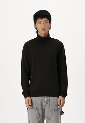 Dunkelgrüner Rollkragenpullover aus weichem Stoff, mit langen Ärmeln und gerippten Bündchen, kombiniert mit hellgrauen Hosen.