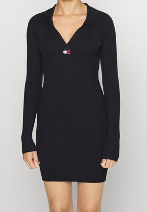 Robe en maille côtelée noire avec manches longues, col en V avec col, et petit logo Tommy Jeans sur la poitrine, portée par une personne debout.