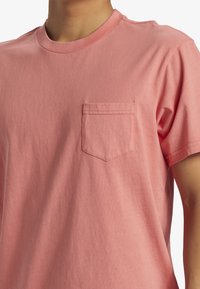 Světle růžové bavlněné tričko s kulatým výstřihem, krátkými rukávy a kapsou na hrudi s rovnou hranou nahoře. Měkká, hladká textura.
