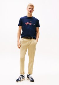 Jūras zila kokvilnas T-kreksa ar "Tommy Jeans" logo, gaišas khaki krāsas šorti un zilas sporta apavus ar baltām akcentiem. Vienkāršs dizains un brīvs griezums.
