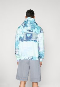 Hanorac cu glugă de culoare albastră deschisă și un model abstract verde, având o buzunar tip „kangaroo” și manșete ribbate. Asortat cu pantaloni scurți gri.