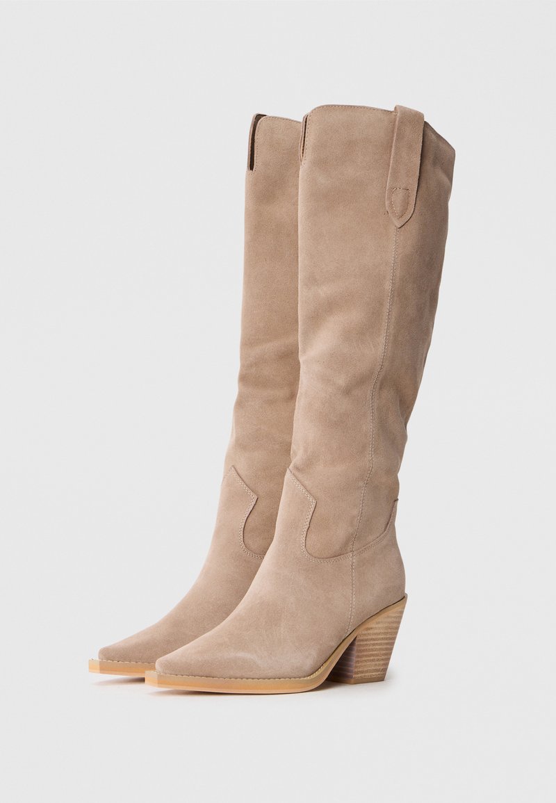 Botas de ante taupe hasta la rodilla, con punteras afiladas y tacón apilado. Presenta costuras laterales y detalles recortados en la parte superior.