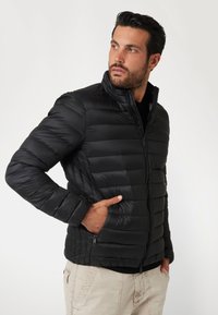 Juodas pūkuotas džemperis su aukštu apykakle, quilted raštu, užtrauktukais kišenėmis ir lygiu paviršiumi, dėvimas virš tamsaus viršaus.