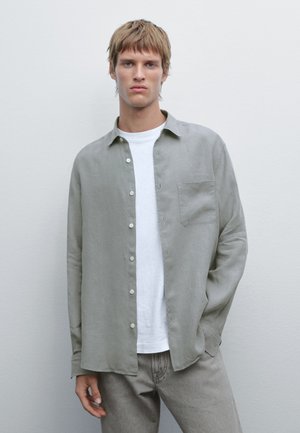 Light gray linen shirt with long sleeves, front button closure, and a patch pocket. Worn over a white T-shirt and paired with light gray pants.