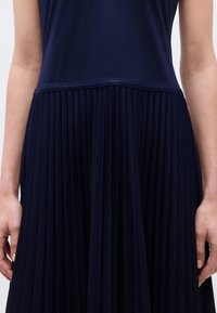 Gros plan sur une personne portant une robe bleu marine avec un haut ajusté et une jupe large plissée, les mains reposant sur les côtés.