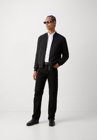Svart bomberjakke med ribbet tekstur, hvit skjorte, svarte jeans og svarte loafers. Tilbehør inkluderer vinklete svarte solbriller.