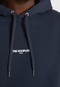 Námořnická modrá mikina s kapucí se stříbrnými průvleky, šňůrkami a bílým logem "THE KOOPLES PARIS" na hrudi. Měkká textilie.