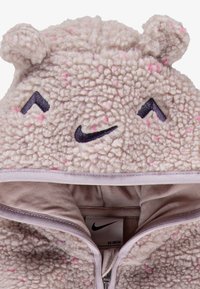 Jasno bež kapuca s ušima medveda, s ružičastim mrljama, nasmejanim licem i crnim Nike logotipom. Mašinski zatvarač s glatkim unutrašnjim podstavljanjem.