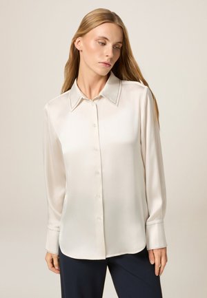 Blusa di seta color crema con colletto classico, chiusura con bottoni sul davanti e maniche lunghe con cuciture a contrasto lungo il colletto e i polsini.
