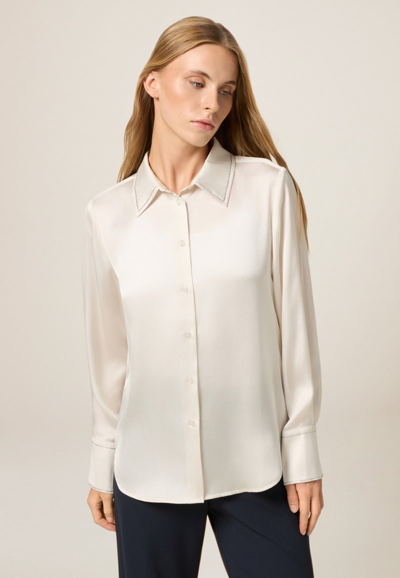 Blusa di seta color crema con colletto classico, chiusura con bottoni sul davanti e maniche lunghe con cuciture a contrasto lungo il colletto e i polsini.