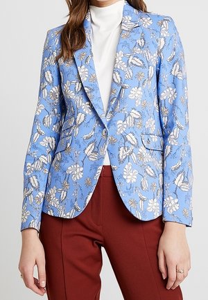Mujer con blazer floral azul claro sobre un jersey de cuello alto blanco, combinado con pantalones de cintura alta color óxido.