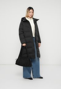 Kvinde iført lang sort puffjakke, cremefarvet halv-zip sweater, vide jeans, sorte sko, mens hun holder en stor sort håndtaske mod en ensfarvet baggrund.