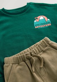 Sweat-shirt vert avec un graphique "AVENTURE EN PLEIN AIR" et un pantalon de survêtement beige avec une taille élastique et un cordon de serrage. Texture de tissu lisse.