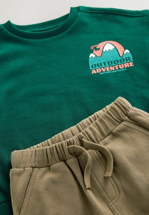 Grøn sweatshirt med "OUTDOOR ADVENTURE" grafik og beige sweatpants med elastisk talje og snøre. Glat stoftekstur.