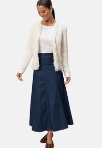 Cardigan en tricot blanc avec des bords à franges, superposé sur un haut blanc ajusté et une longue jupe en jean foncé, associé à des chaussures marron à clous.