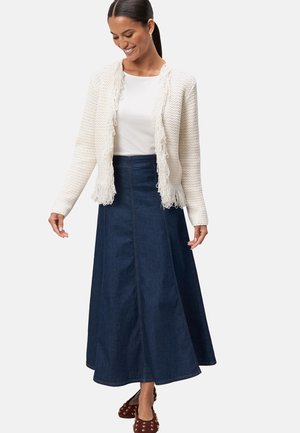 Cardigan en tricot blanc avec des bords à franges, superposé sur un haut blanc ajusté et une longue jupe en jean foncé, associé à des chaussures marron à clous.