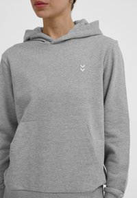 Felpa con cappuccio grigia in materiale morbido, con tasca frontale, polsini a costine e un piccolo logo a chevron bianco sul petto.