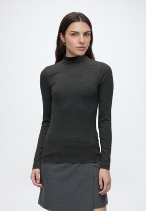 Vrouw met lang donker haar, kleine creolen, een donkergrijze coltrui en een grijze rok tegen een effen witte achtergrond.
